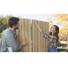 Ein Paar montiert gemeinsam im Freien ein vertikales Lamellen Sichtschutzelement aus Holz für den Garten.