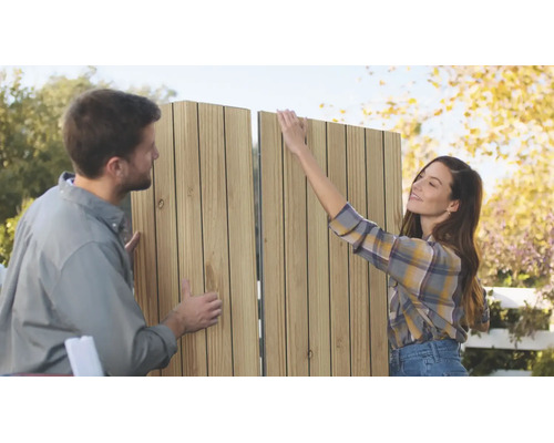 Ein Paar montiert gemeinsam im Freien ein vertikales Lamellen Sichtschutzelement aus Holz für den Garten.