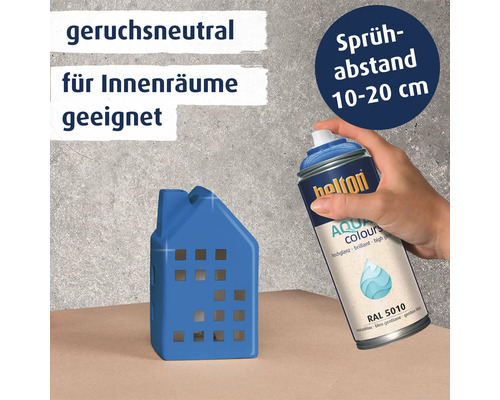 Eine Hand sprüht mit einer Hellon Aqua Colours Sprühdose ein kleines Hausmodell an, mit Hinweisen zu Geruchsneutralität und Sprühabstand.