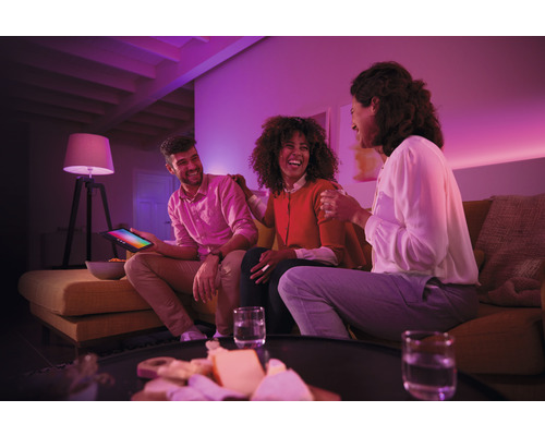 Drei Personen sitzen in einem Wohnzimmer mit farbiger Beleuchtung auf einer Couch und lachen.