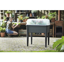 Hochbeet mit transparentem Deckel im Garten, mit bepflanzten Behältern im Hintergrund.