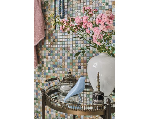 Dekoratives Arrangement mit Blumen, Vogel, Teekanne und Flakon auf einem Beistelltisch vor einer Mosaikwand