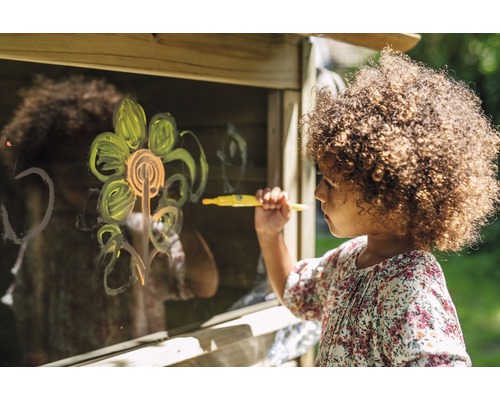 Ein Kind malt mit einem Stift ein Bild auf eine Glasscheibe eines Holzspielhauses.