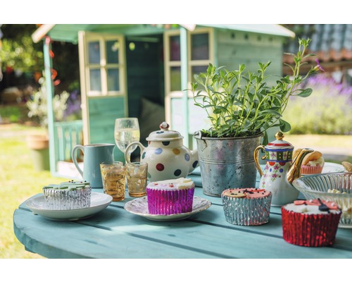 Gedeckter Tisch mit Gartenhaus im Hintergrund für eine Gartenparty mit Teekannen, Pflanzen, Gläsern und Muffins.