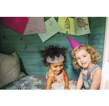 Zwei Mädchen mit Partyhüten und Kinderzeichnungen an der Holzwand im Spielhaus.