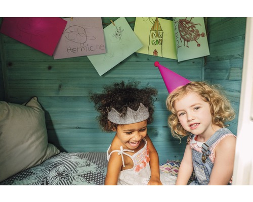 Zwei Mädchen mit Partyhüten und Kinderzeichnungen an der Holzwand im Spielhaus.