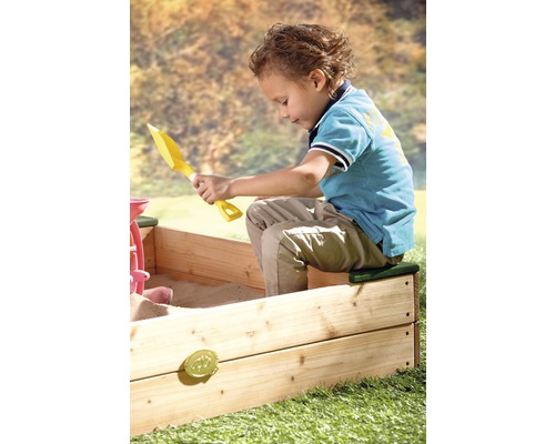 Junge spielt mit einem gelben Spaten in einem Sandkasten aus Holz.