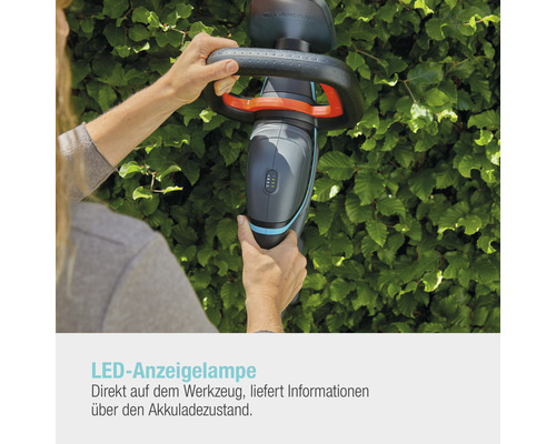 Person bedient eine Akku Heckenschere der Marke Gardena vor einer grünen Hecke.