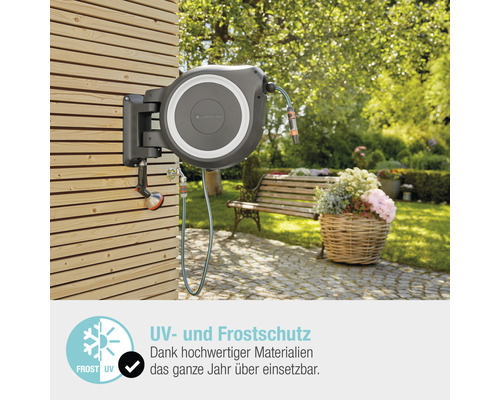 Auf einer Holzwand montierte Gardena Schlauchbox mit Gartenschlauch und Sprühkopf vor Gartenkulisse mit Bank und Blumenkorb.