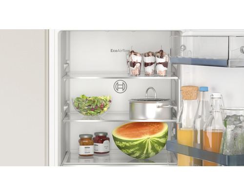 Innenansicht eines Kühlschranks mit verschiedenen Lebensmitteln wie Desserts, Salat, Kochtopf, Wassermelone und Getränken.