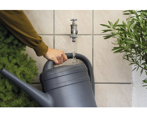Eine Person füllt eine graue Gießkanne mit Wasser aus einem Wasserhahn im Garten.