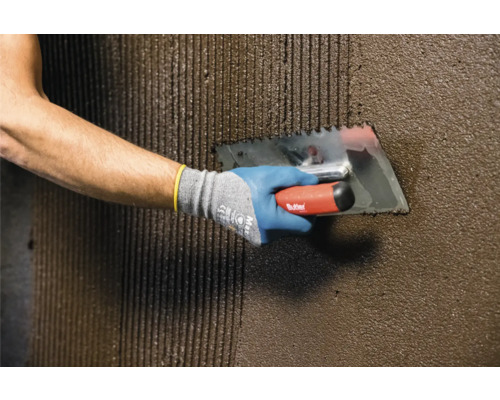 Handwerker trägt mit einer Zahnspachtel Kleber auf eine Wand auf.