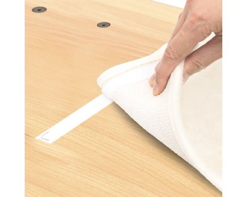 Eine Person befestigt einen Teppich mit einem Klebestreifen auf einem Holzboden.