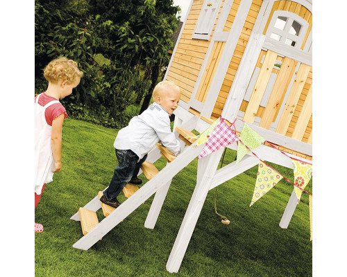 Kind klettert auf ein Holzspielhaus im Garten, dekoriert mit einer bunten Wimpelkette.