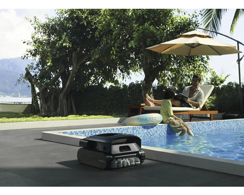 Szene mit einem Poolreinigungsroboter neben einem Schwimmbad, in dem sich Personen unter einem Sonnenschirm entspannen.