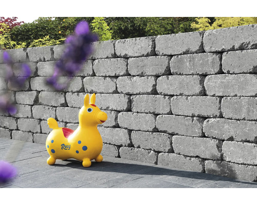 Gestapelte graue Betonsteine bilden eine Gartenmauer mit einem Kinderspielzeug davor.