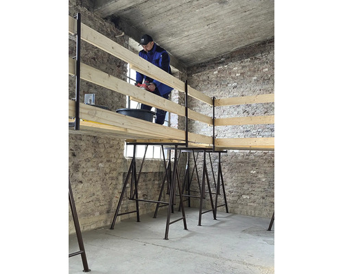 Ein Mann arbeitet mit einer Bohrmaschine an einer Holzkonstruktion, die auf Gerüstböcken steht.