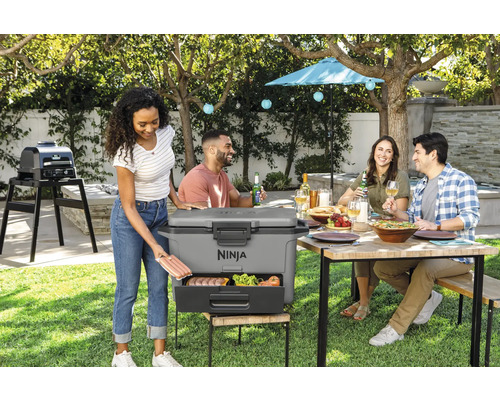 Szene im Freien mit einer Gruppe von Menschen, einem tragbaren Ninja-Grill mit herausziehbarer Schublade, in der Lebensmittel gelagert werden, und einem Tisch mit Essen und Getränken
