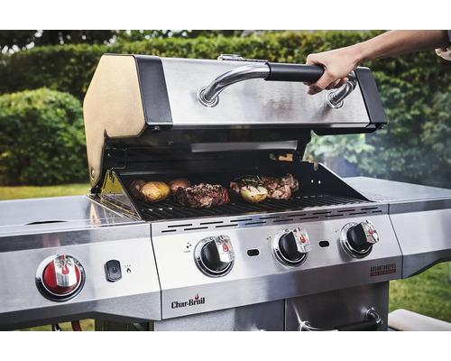 Person öffnet den Deckel eines Edelstahl-Gasgrills mit Steaks und Kartoffeln auf dem Grillrost.