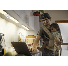 Ein Mann mit Babytrage bedient einen Laptop in einer Küche mit Unterbauleuchten.