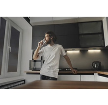 Mann trinkt ein Glas Wasser in einer Küche mit Mikrowelle, Kochfeld und Wasserkocher.