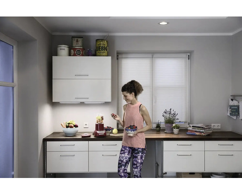 Helle Küche mit einer Frau, die Obst für einen Smoothie zubereitet. Ein Mixer steht auf der Küchentheke.