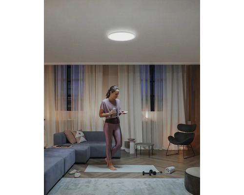 Eine Frau steht in einem Wohnzimmer mit einer runden Deckenleuchte, einem Sofa und einem Stuhl.