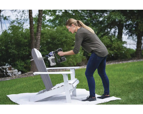 Eine Frau sprüht mit einer Farbspritzpistole Farbe auf einen Adirondack-Stuhl im Freien.
