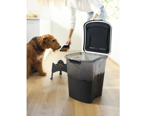 Eine Person füllt Tiernahrung mit einer Schaufel in einen Hundenapf, neben einem Behälter für Tiernahrung und einem Hund