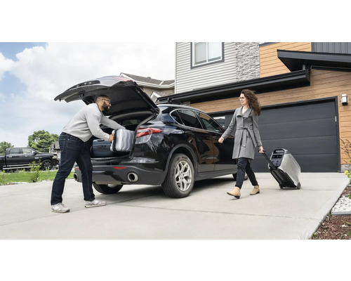 Ein Mann packt Koffer in den Kofferraum eines Autos, während eine Frau einen tragbaren Stromgenerator auf Rädern zieht.