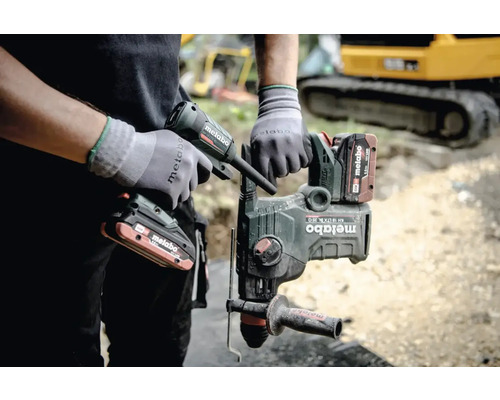 Ein Mann hält einen Metabo Bohrhammer und eine Kartuschenpistole in der Hand, im Hintergrund ist ein Bagger zu sehen