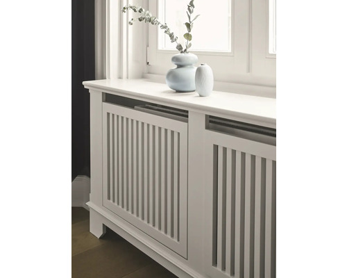 Heizkörperverkleidung aus Holz mit vertikalen Lamellen und zwei Vasen auf der Oberseite.