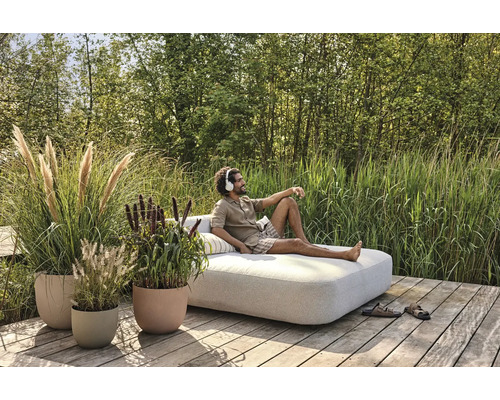 Mann mit Kopfhörern entspannt auf einer Gartenliege, umgeben von dekorativen Pflanzgefäßen und üppigem Grün.