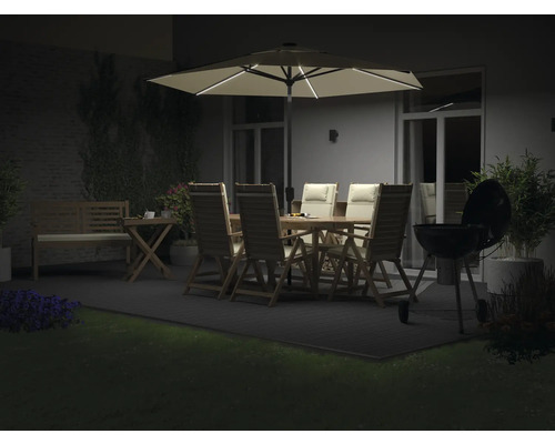 Gartenambiente mit beleuchtetem Sonnenschirm, Gartentisch, Stühlen, Grill und Bepflanzung bei Nacht.