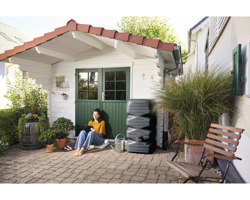 Gartenszene mit einer Frau, die neben einem Regenwassertank vor einem Gartenhaus sitzt