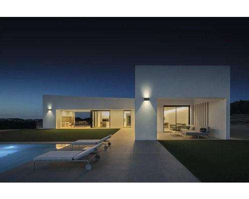 Moderne Hausfassade mit Außenbeleuchtung, Pool und Liegestühlen in der Abenddämmerung.