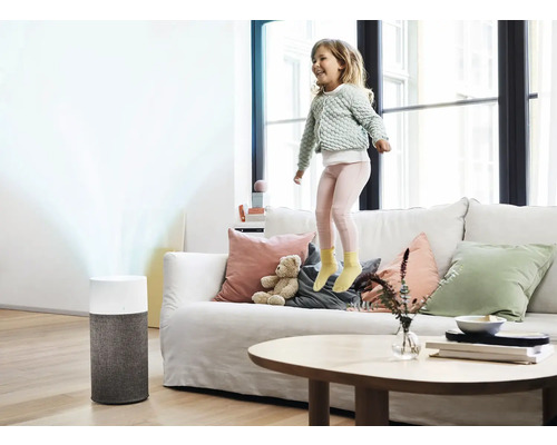 Ein Mädchen springt auf einem Sofa in einem hellen Wohnzimmer, im Hintergrund steht ein Luftreiniger