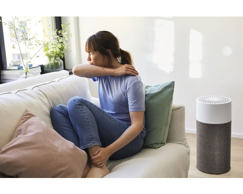 Eine Frau hustet auf einem Sofa neben einem Luftreiniger.