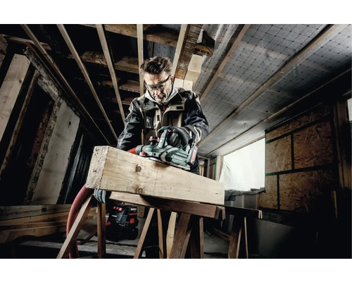 Handwerker mit Schutzbrille hobelt ein Holzbrett mit einem Elektrohobel von Metabo in einem Rohbau.
