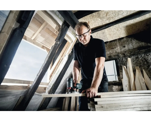 Ein Mann arbeitet mit einer Oberfräse an einem Holzbrett in einem Dachgeschoss.