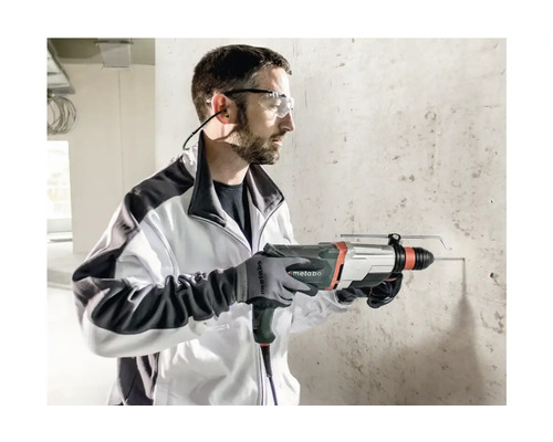 Mann bohrt mit Bohrhammer in Betonwand. Er trägt Schutzbrille, Gehörschutz und Handschuhe.