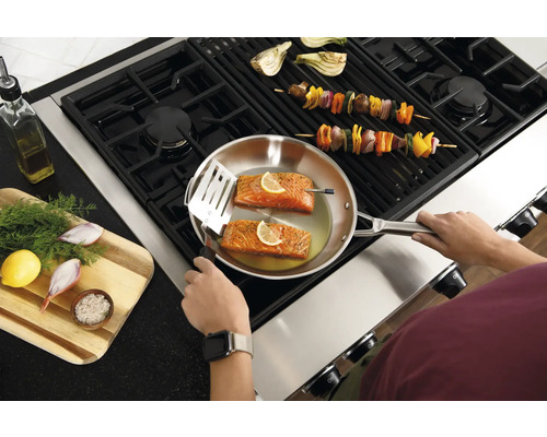 Eine Person kocht Lachs in einer Pfanne auf einem Herd, während Gemüsespieße auf dem Grill liegen.