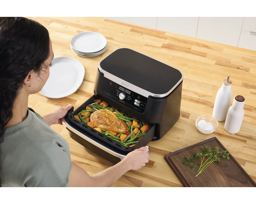 Eine Frau schiebt eine Heißluftfritteuse mit Hähnchen und Gemüse auf einem Holztisch zusammen mit Tellern, Ölflaschen und Kräutern zusammen.