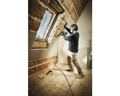Ein Mann befestigt eine Gipskartonplatte mit einem Druckluftnagler in einem Dachgeschossraum mit Dachfenster und Holzfußboden. Er trägt dabei eine Atemschutzmaske und Gehörschutz.