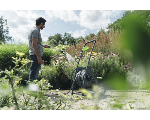 Ein Mann bewässert mit einem Gartenschlauch Blumen, der Schlauch ist auf einer Schlauchwagen aufgerollt.