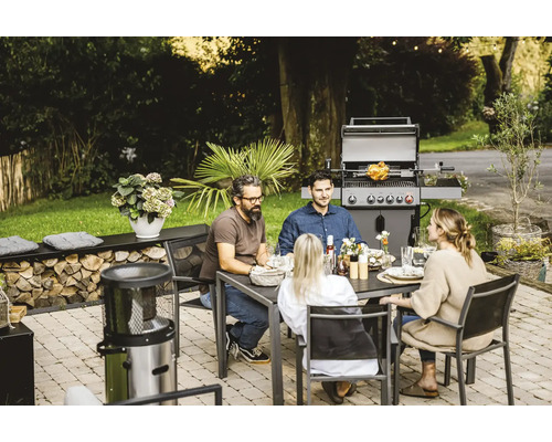 Personen sitzen an einem Gartentisch mit Grill im Hintergrund.