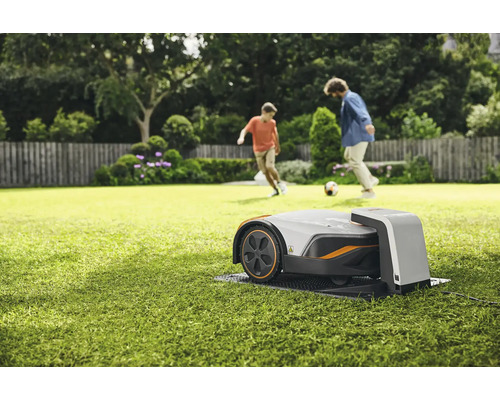 Rasenmähroboter auf Rasen mit spielenden Personen im Hintergrund