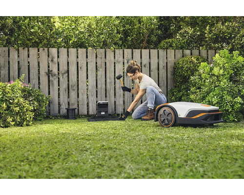 Frau installiert einen Mähroboter mit Hammer und Begrenzungskabel im Garten.