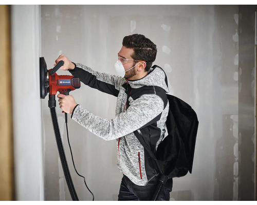 Ein Mann schleift eine Wand mit einem Einhell Trockenbauschleifer, Staubmaske und Schutzbrille sind getragen.