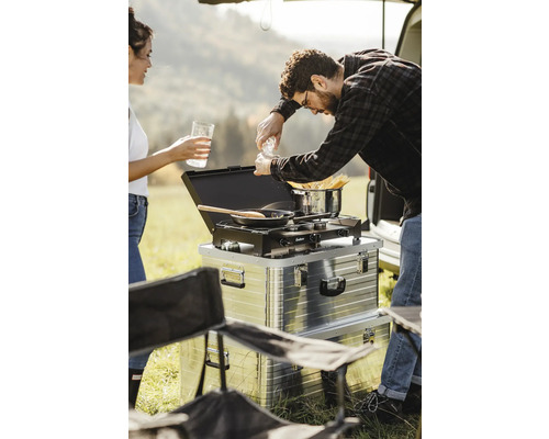 Campingküche mit Kocher, Pfanne und Topf im Outdoorbereich, zwei Personen kochen.
